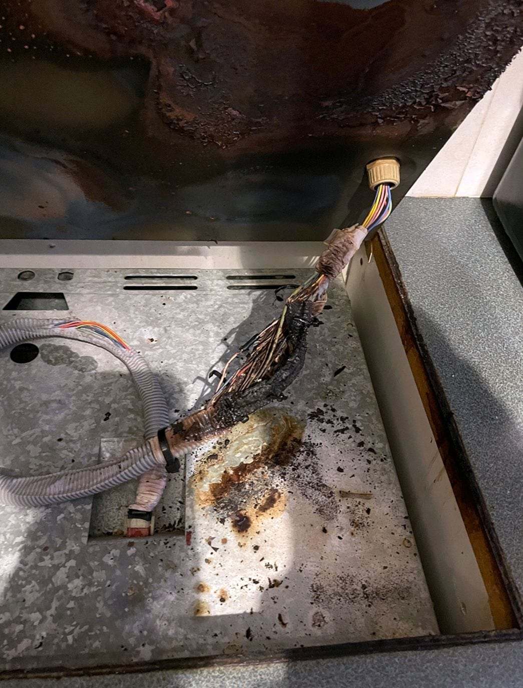 Scorched and melted wiring harness where the cable enters a metal appliance cabinet, soot on the panel beside it