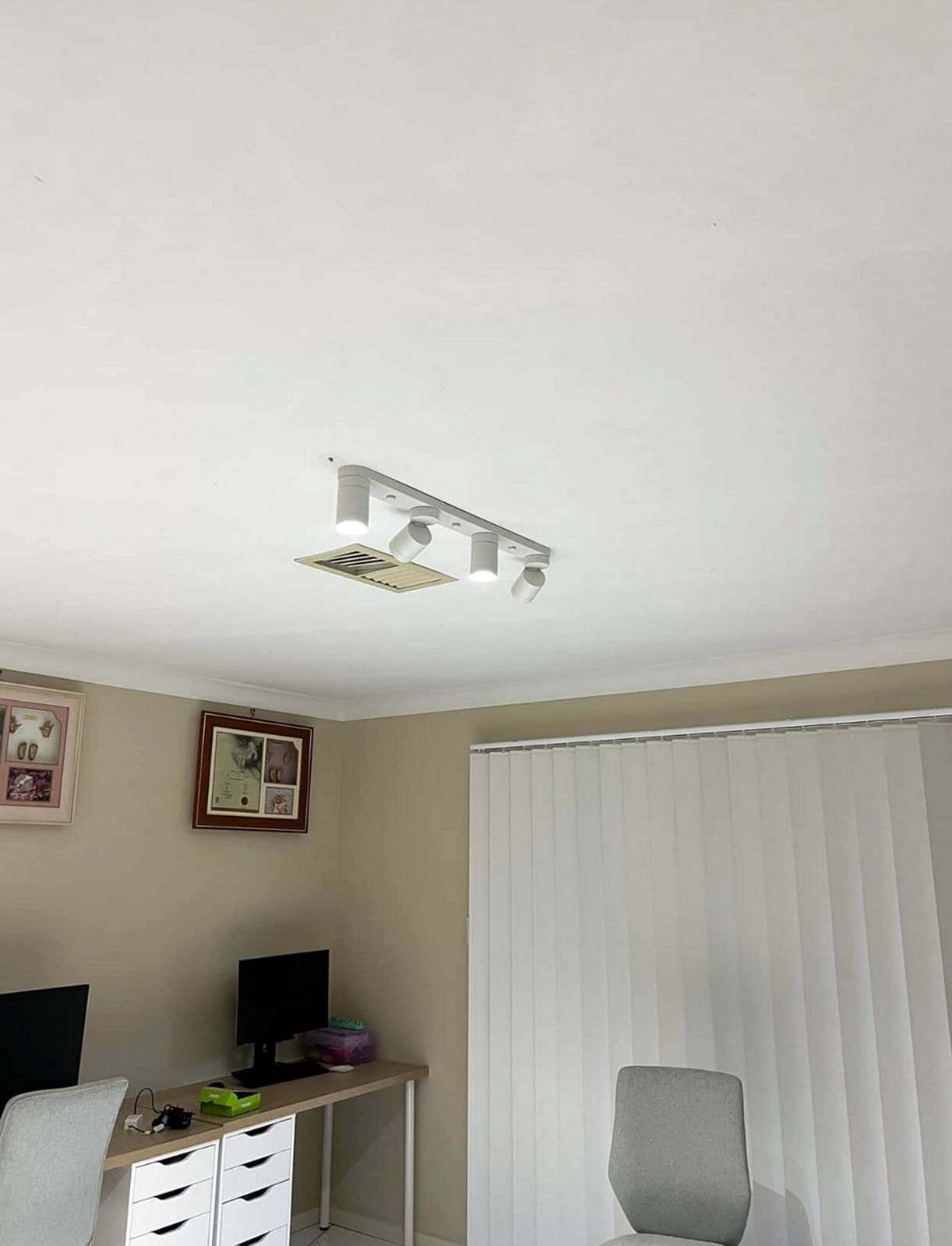 White four-head track light mounted on the ceiling of a home office above a desk and a window with vertical blinds