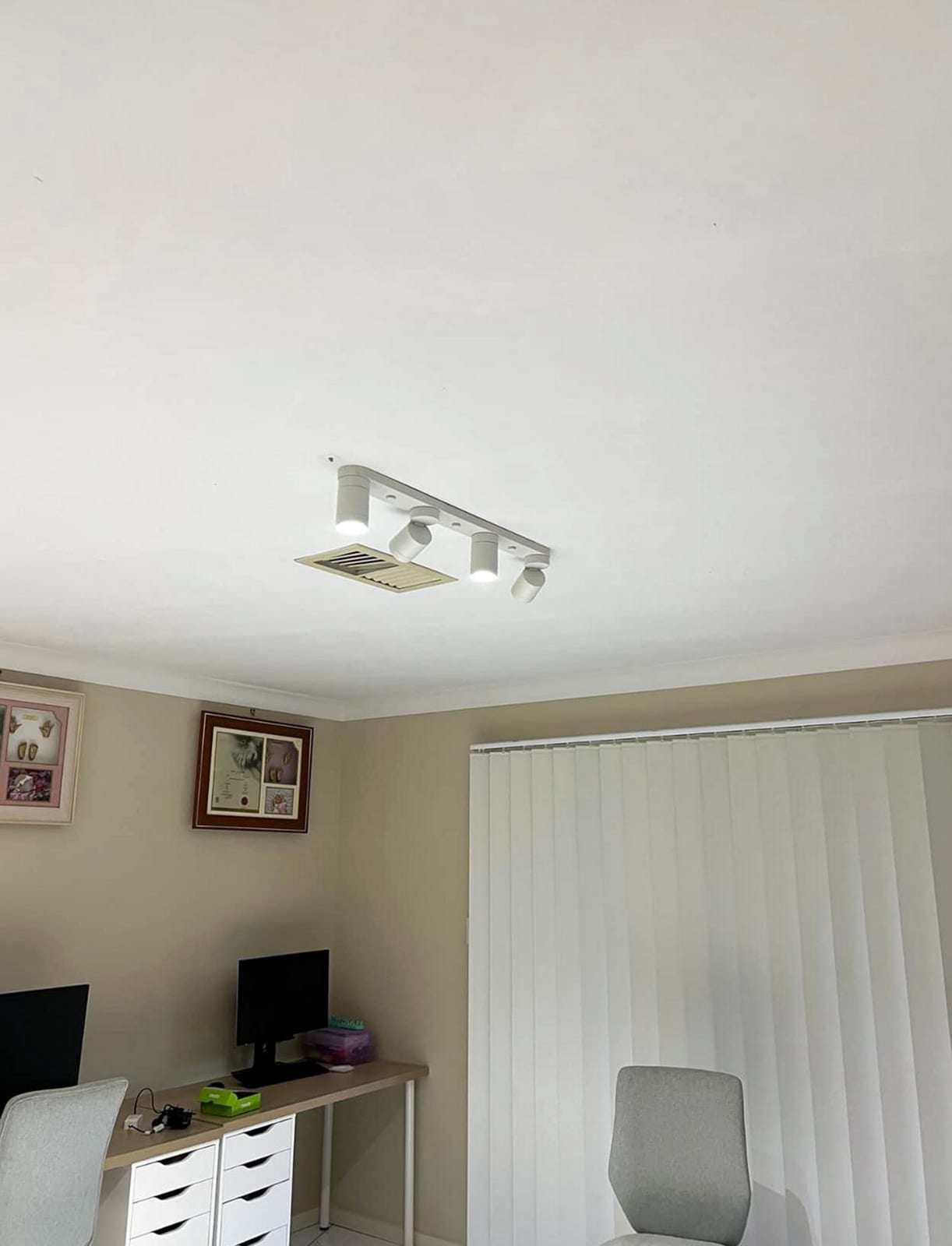 White four-head track light mounted on the ceiling of a home office above a desk and a window with vertical blinds