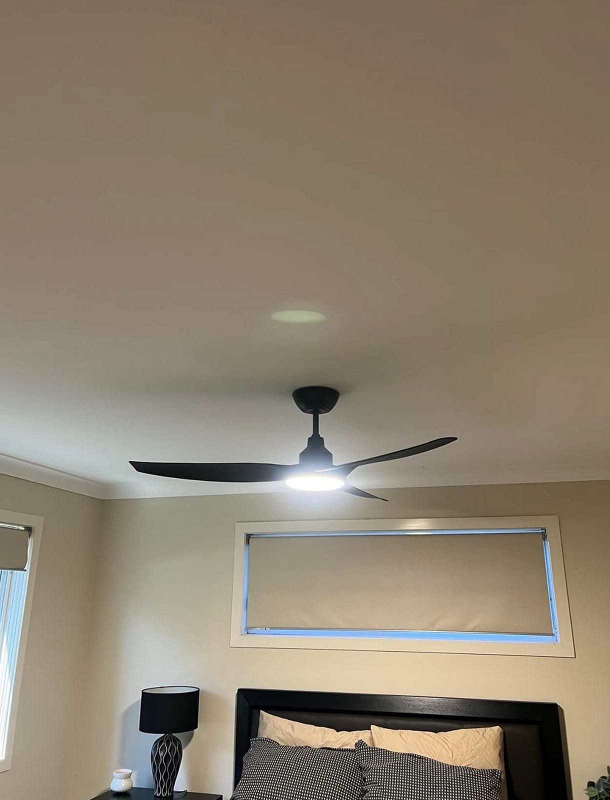 Black three-blade ceiling fan with its LED light switched on above a bed with a dark headboard
