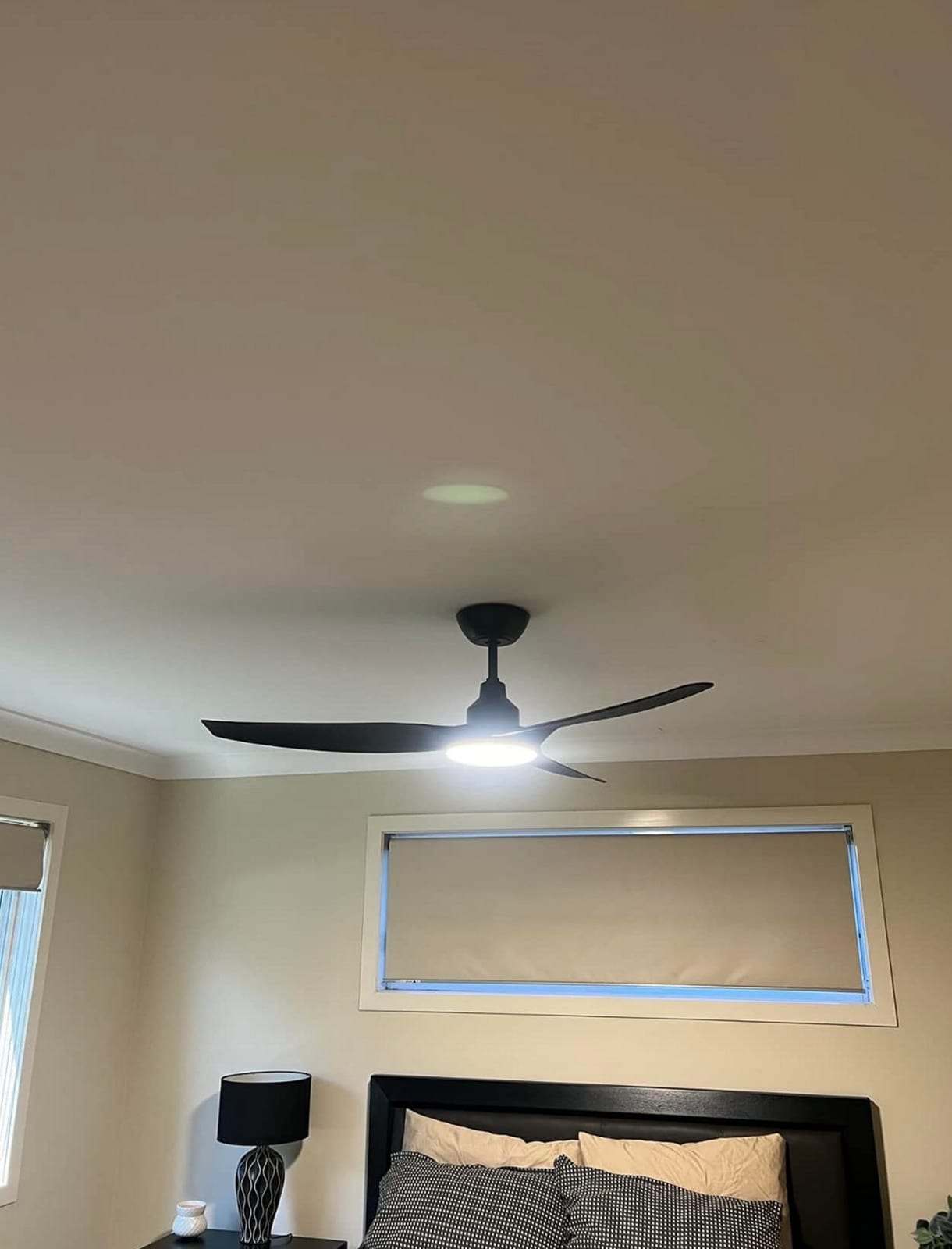 Black three-blade ceiling fan with its LED light switched on above a bed with a dark headboard