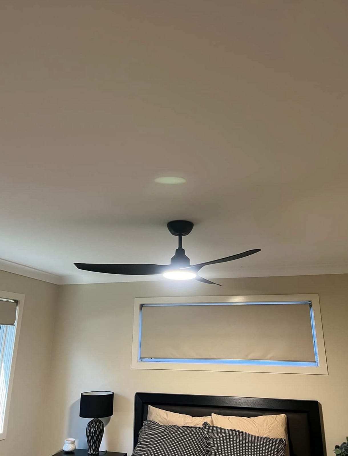 Black three-blade ceiling fan with its LED light switched on above a bed with a dark headboard