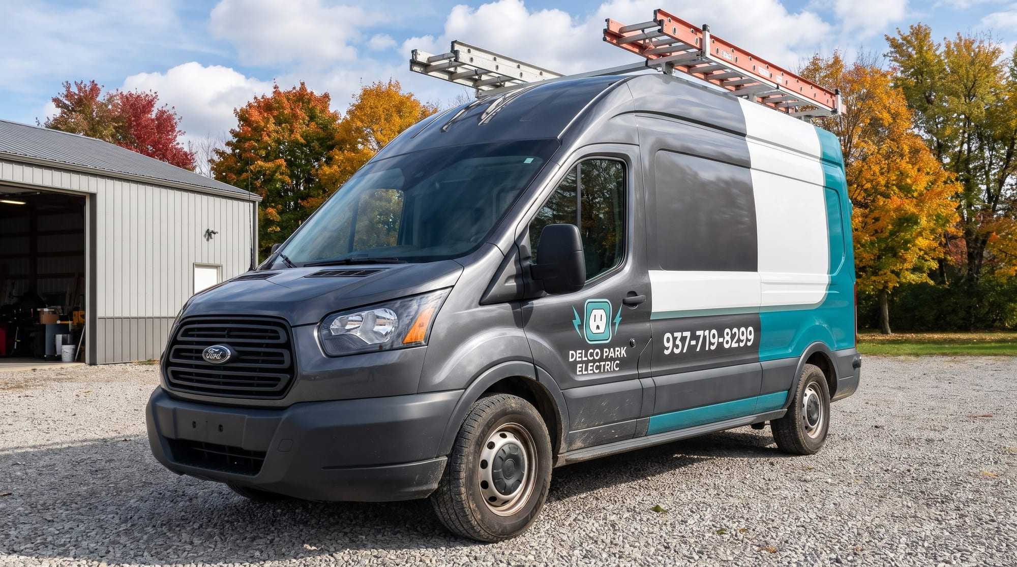 Dark gray, white and teal Ford Transit van with the Delco Park Electric logo on its front door and the phone number on its side, parked on gravel before autumn maples